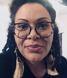 a light golden brown skinned femme with grey-brown locs and glasses smiles softly at the camera.