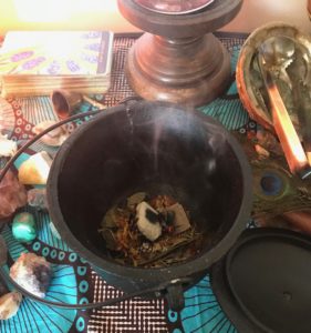 a black cauldron with burning herbs sits among crystals and tarot cards on an altar draped in turquoise ankara fabric.