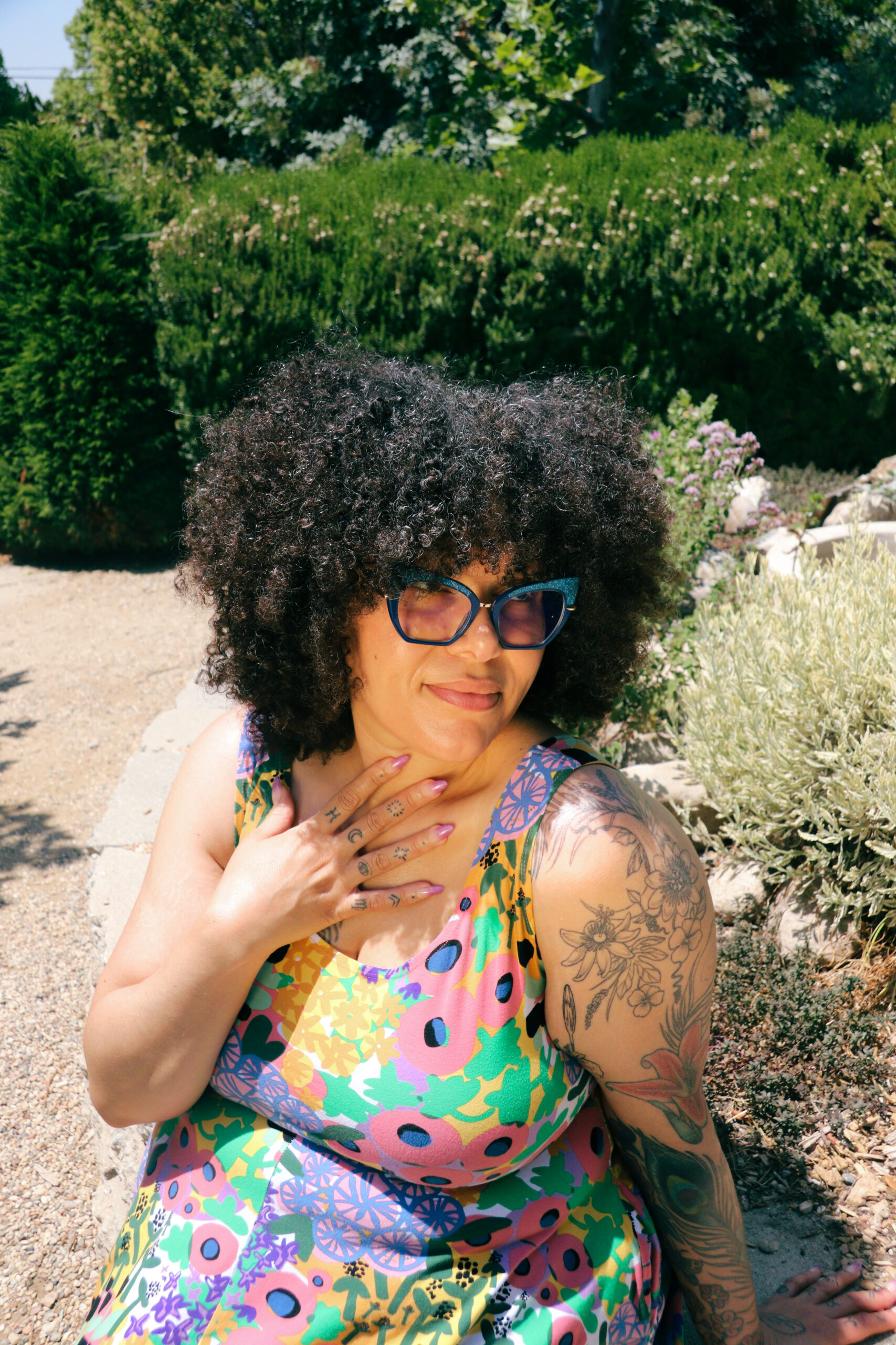 Tasha Fierce, a very light brown skinned mixed black femme with mid-length curly hair and tattoos, wearing cat-eye glasses and a brightly patterned dress. They sit in front of some bushes with their hand over their heart and are looking off to the side.